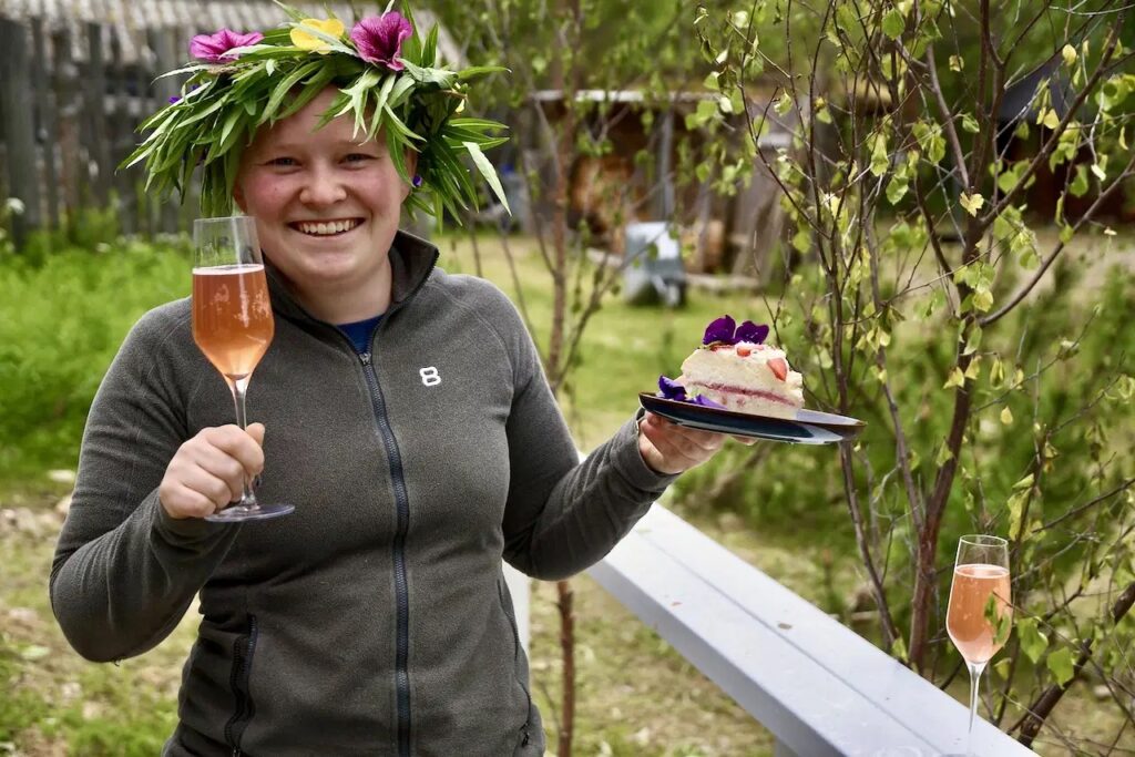 Hymyilevä henkilö, jolla on kukkakruunu päällään, pitelee lasillista vaaleanpunaista juomaa ja kakkulautasta ulkona, vehreyden ja puiden ympäröimänä. Toinen lasi vaaleanpunaista juomaa istuu läheisellä kaiteella.