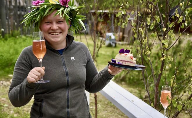 Hymyilevä henkilö, jolla on kukkakruunu päällään, pitelee lasillista vaaleanpunaista juomaa ja kakkulautasta ulkona, vehreyden ja puiden ympäröimänä. Toinen lasi vaaleanpunaista juomaa istuu läheisellä kaiteella.