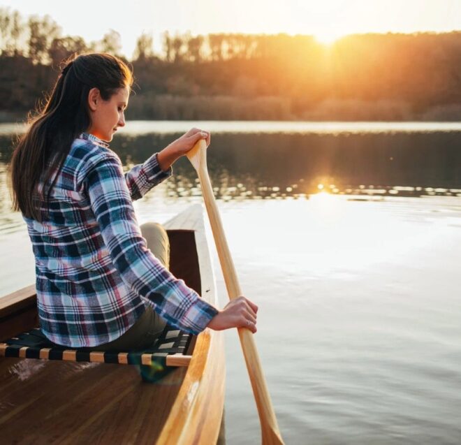Ruudulliseen paitaan pukeutunut nainen meloo puukanoottia rauhallisella järvellä auringonlaskun aikaan, puiden ympäröimänä ja lempeän auringonvalon heijastuessa veteen.