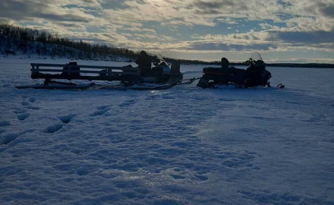 Lumipeitteinen maisema osittain pilvisellä taivaalla, jonka läpi paistaa aurinko. Kaksi moottorikelkkaa tai -kelkkaa on pysäköity lumeen, ja jäljet johtavat niitä kohti. Puut reunustavat horisonttia kaukana.