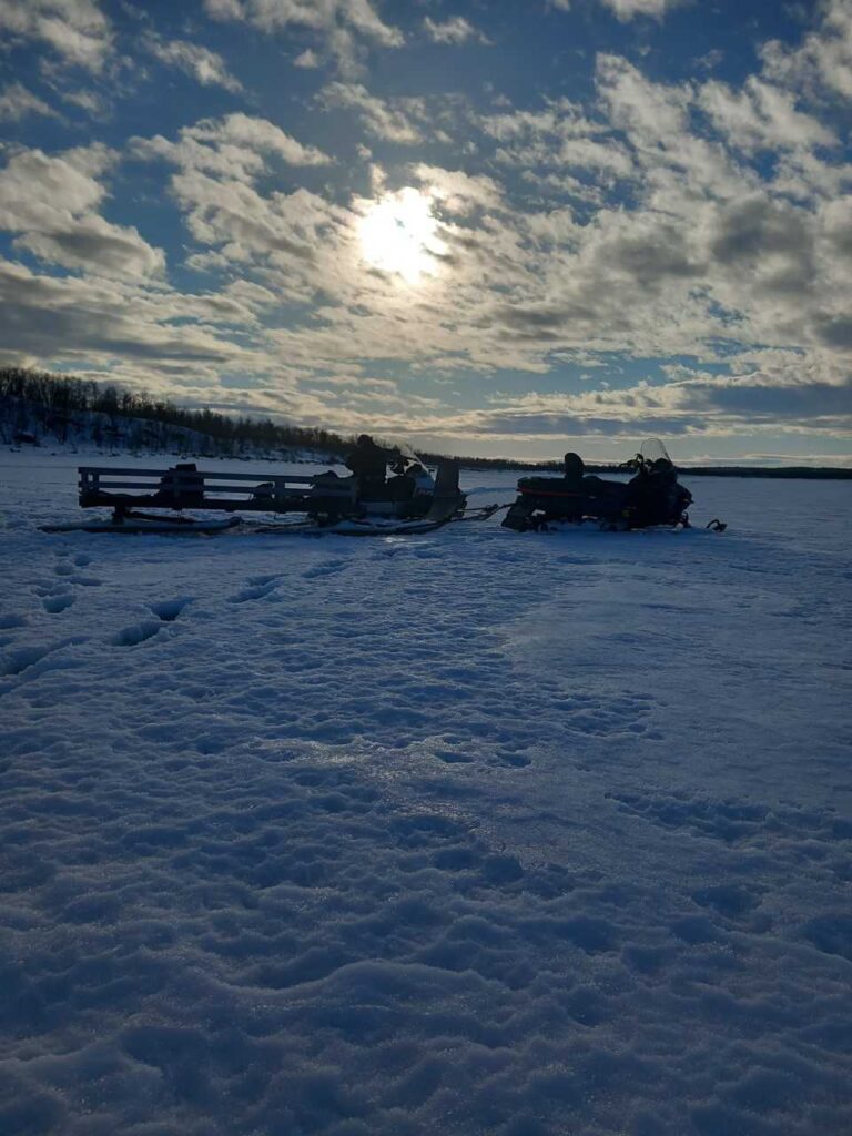 Lumipeitteinen maisema osittain pilvisellä taivaalla, jonka läpi paistaa aurinko. Kaksi moottorikelkkaa tai -kelkkaa on pysäköity lumeen, ja jäljet johtavat niitä kohti. Puut reunustavat horisonttia kaukana.
