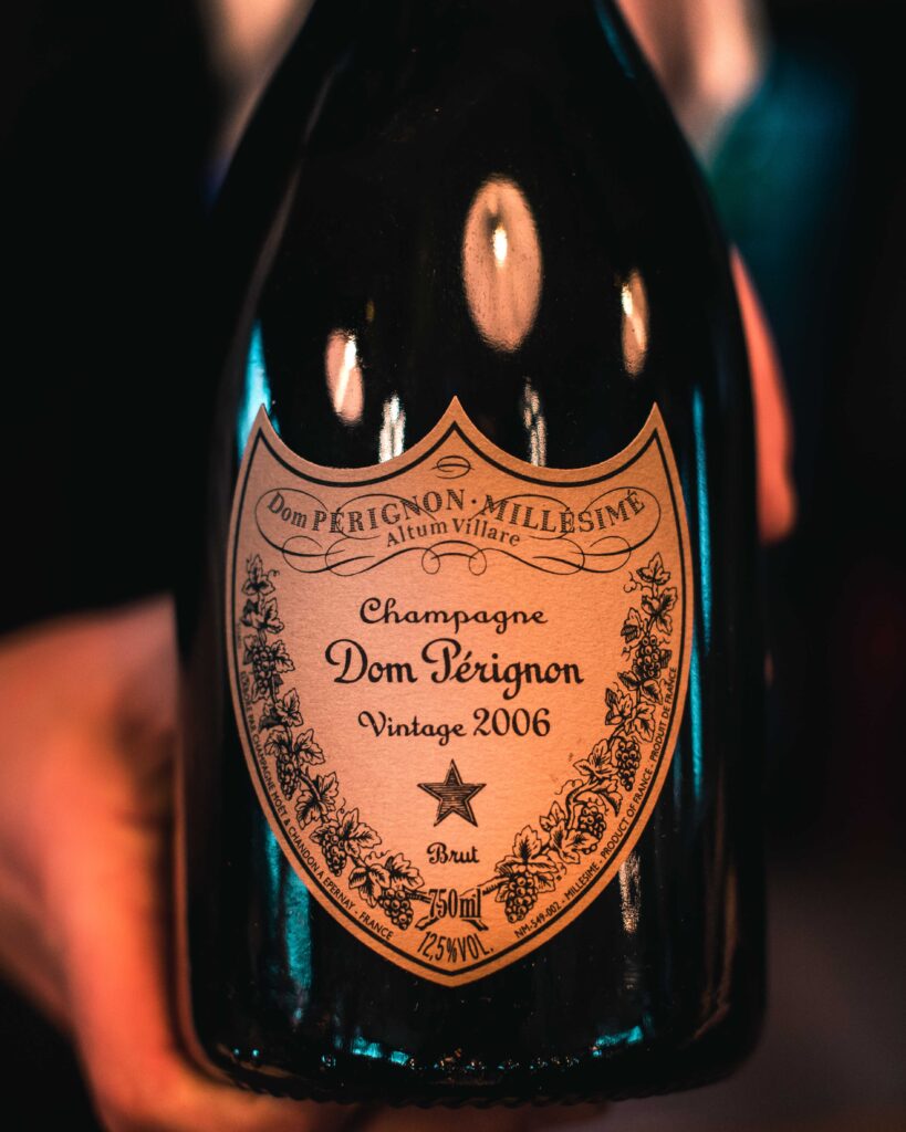Lähikuva kädestä, jossa on Dom Pérignon Vintage 2006 -samppanjapullo, jossa näkyy koristeltu etiketti, jossa on kultaisia ja mustia yksityiskohtia ja teksti selvästi näkyvissä.
