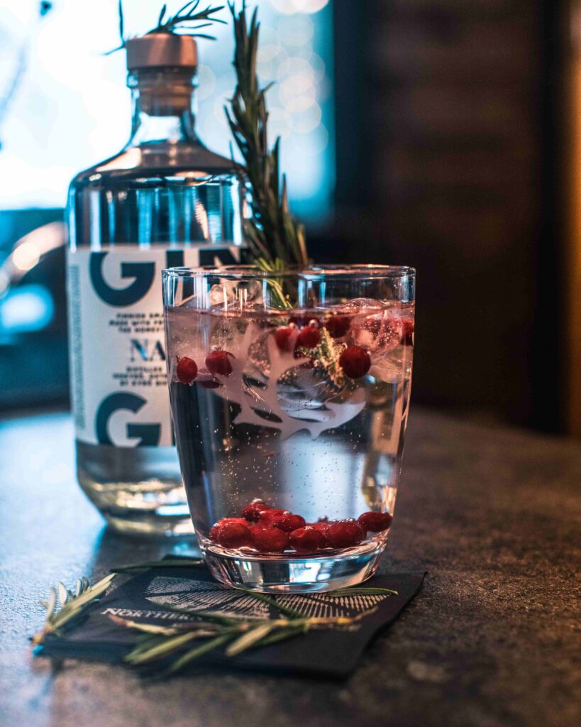 Lasi gin tonicia jäillä, granaattiomenan siemenillä ja rosmariininoksalla on lautasliinan päällä ginipullon vieressä tummalla työtasolla.