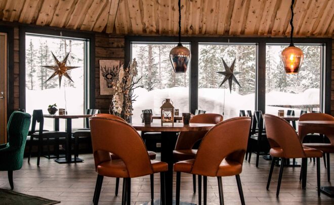 Viihtyisä ravintola, jossa on puulattiat ja puinen katto, ruskeat nahkatuolit pöytien ympärillä, suuret ikkunat, joista näkyy luminen maisema, ja katosta roikkuvat lämpimät riippuvalaisimet.