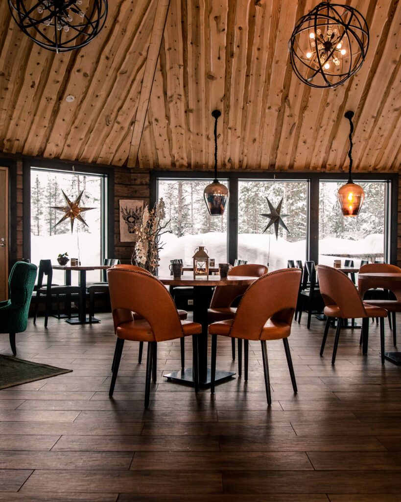Viihtyisä ravintola, jossa on puulattiat ja puinen katto, ruskeat nahkatuolit pöytien ympärillä, suuret ikkunat, joista näkyy luminen maisema, ja katosta roikkuvat lämpimät riippuvalaisimet.