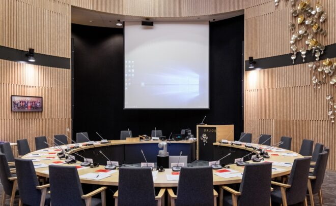 Nykyaikainen kokoushuone, jossa on suuri pyöreä pöytä, siististi asetellut tuolit, paperimateriaalia, keskellä mikrofoni ja edessä valkokangas. Huoneessa on puupaneeliseinät ja koristeellinen valaistus oikealla.