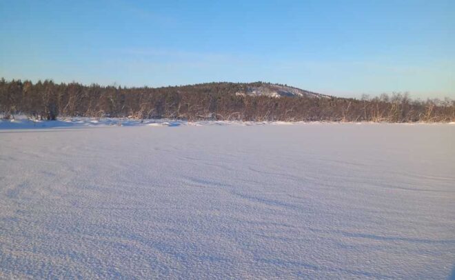 Laaja, lumen peittämä pelto levittäytyy kaukaisuuteen kirkkaansinisen taivaan alla, ja horisontissa näkyy puurivi ja pieni kukkula.