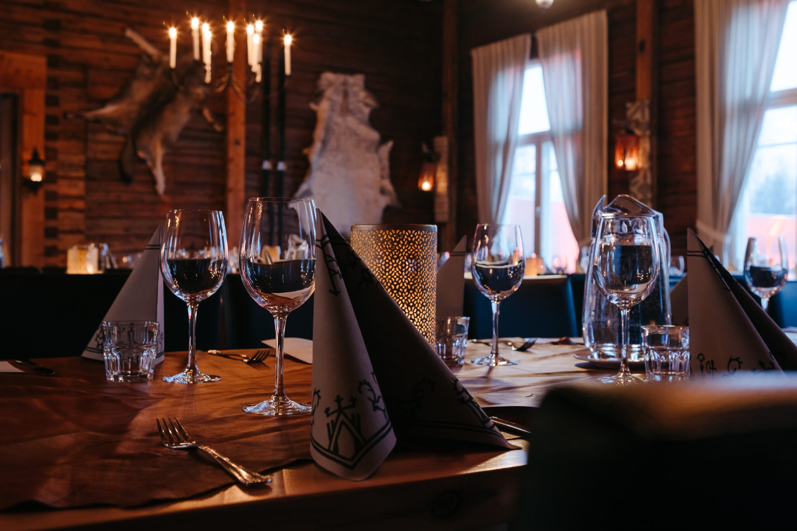 Hämärästi valaistu, maalaismainen ravintola, jonka seinät ovat puuta ja jossa on katetut pöydät, joissa on taitettuja lautasliinoja, viinilaseja ja vesikannuja. Suuret ikkunat, joissa on valkoiset verhot, päästävät sisään pehmeää päivänvaloa. Kruunukruunut ja kynttilät lisäävät viihtyisää tunnelmaa.