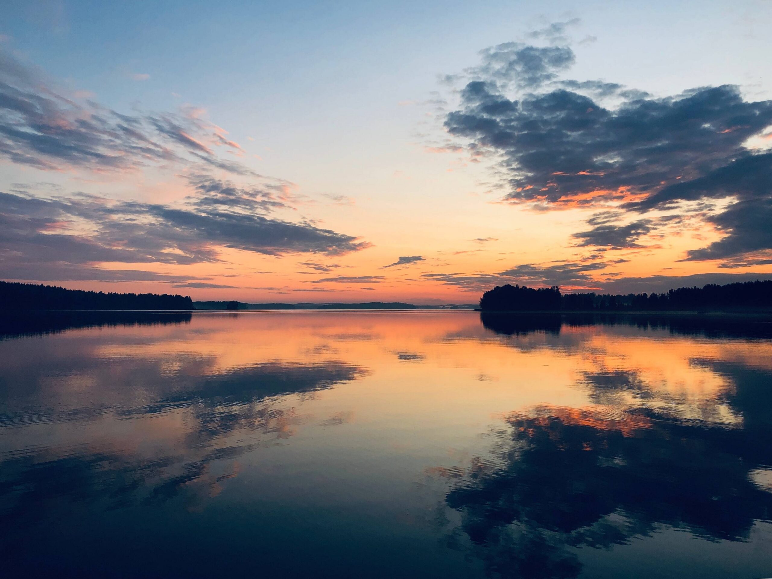 Rauhallinen järvi auringonlaskun aikaan, taivas on oranssin, vaaleanpunaisen ja sinisen sävyinen, pilvet peittävät osittain auringon ja veden pinnalla heijastuu taivas ja puut.