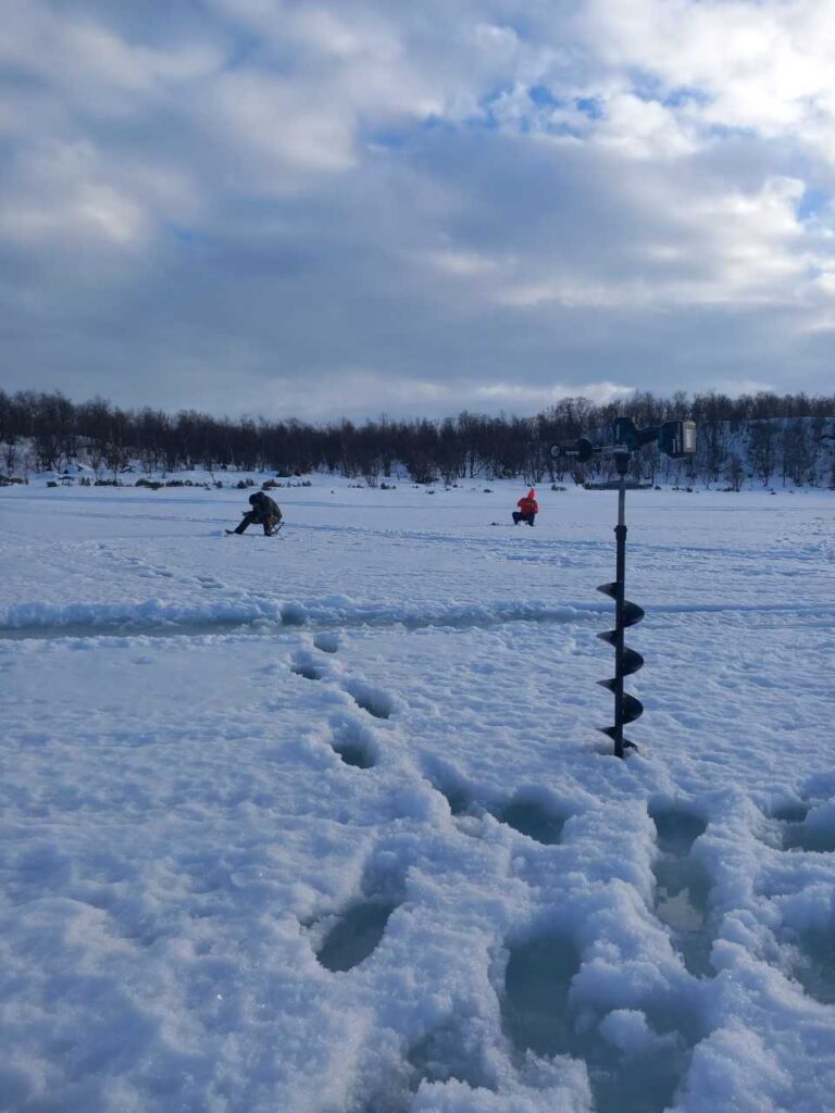 Kaksi ihmistä istuu lumisella, jäätyneellä järvellä jääkalastamassa pilvisen taivaan alla. Etualalla lumeen on juuttunut jääpora, ja jalanjäljet johtavat kohti taustalla olevia ihmisiä. Horisonttia reunustavat puut.