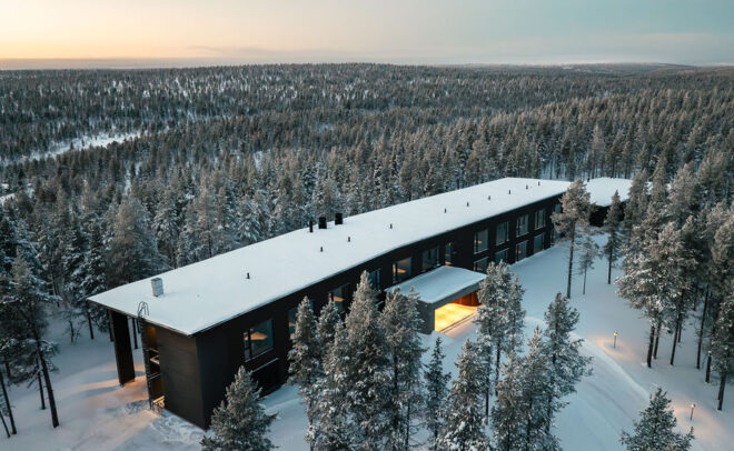 Modernia, suorakulmaista rakennusta, jonka katto on lumipeitteinen, ympäröi auringonlaskun aikaan tiheä luminen mäntymetsä, joka luo seesteisen talvimaiseman. Lämpimät valot loistavat rakennuksen osista.