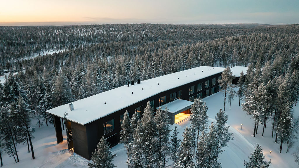Modernia, suorakulmaista rakennusta, jonka katto on lumipeitteinen, ympäröi auringonlaskun aikaan tiheä luminen mäntymetsä, joka luo seesteisen talvimaiseman. Lämpimät valot loistavat rakennuksen osista.