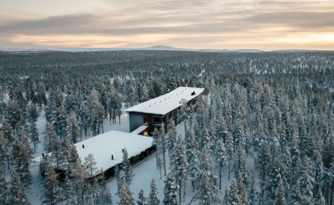 Ilmakuva modernista rakennuksesta, jossa on lumipeitteiset katot ja jota ympäröi tiheä metsä lumisia puita pilvisen taivaan alla auringonnousun tai -laskun aikaan.