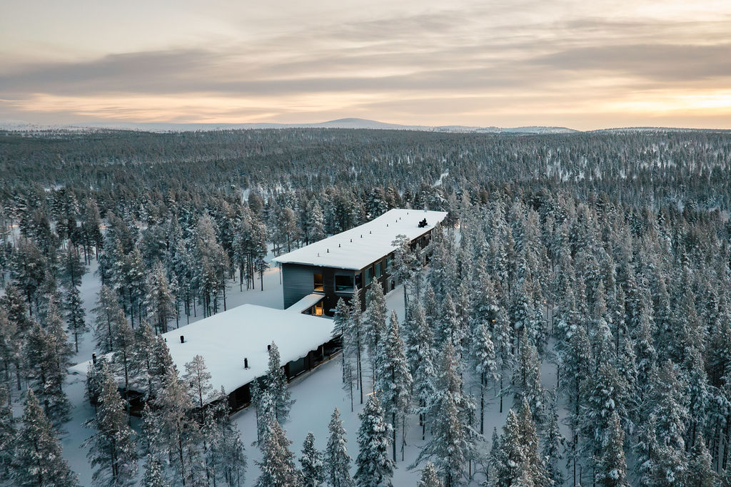 Ilmakuva modernista rakennuksesta, jossa on lumipeitteiset katot ja jota ympäröi tiheä metsä lumisia puita pilvisen taivaan alla auringonnousun tai -laskun aikaan.