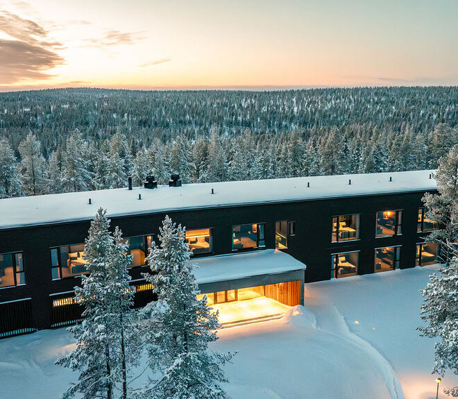 Modernia suorakulmaista rakennusta, jossa on suuret ikkunat, ympäröivät lumiset puut talvimaisemassa, lämpimät valot loistavat sisällä ja luminen metsä ulottuu auringonlaskun aikaan horisonttiin.