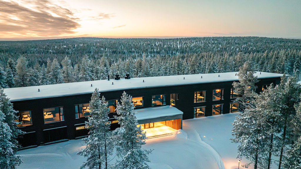 Modernia suorakulmaista rakennusta, jossa on suuret ikkunat, ympäröivät lumiset puut talvimaisemassa, lämpimät valot loistavat sisällä ja luminen metsä ulottuu auringonlaskun aikaan horisonttiin.