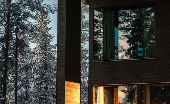 Moderni puinen mökki, jossa on suuret ikkunat, loistaa lämpimästi lumisessa metsässä iltahämärässä, heijastaen ympäröiviä lumipeitteisiä puita.