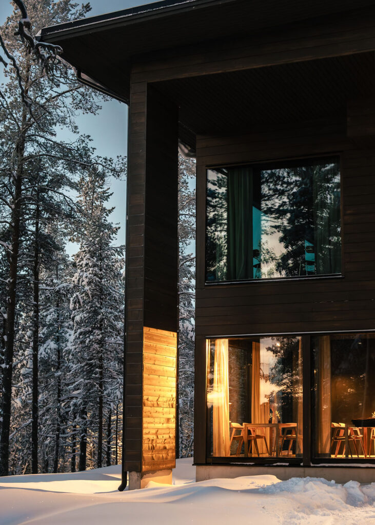 Moderni puinen mökki, jossa on suuret ikkunat, loistaa lämpimästi lumisessa metsässä iltahämärässä, heijastaen ympäröiviä lumipeitteisiä puita.