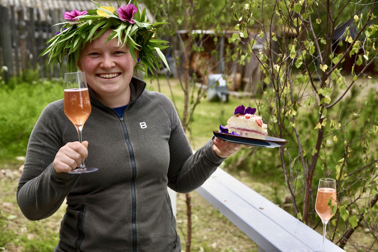 Hymyilevä henkilö, jolla on kukkakruunu päällään, pitelee lasillista vaaleanpunaista juomaa ja lautasella koristeltua kakunpalaa ulkona, vehreyden ympäröimänä. Toinen lasi vaaleanpunaista juomaa istuu läheisellä valkoisella kaiteella.