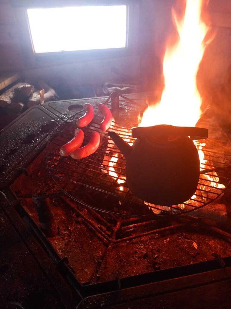 Kolme makkaraa ja musta vedenkeitin paistuvat metalliritilällä avotulen yllä, liekit nousevat korkealle; taustalla näkyy pieni ikkuna, joka päästää luonnonvaloa sisään.