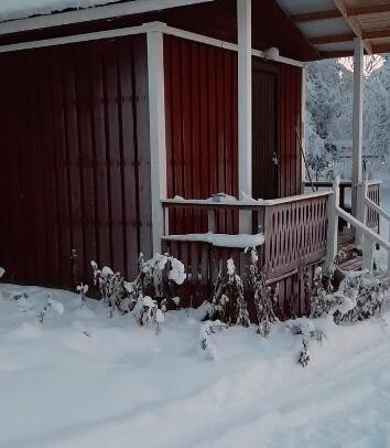 Pieni punainen puinen mökki, jossa on valkoiset reunukset, seisoo lumisessa maisemassa. Lumi peittää katon, portaat ja maan, ja kuistin kaiteen lähellä näkyy paljaita kasveja.