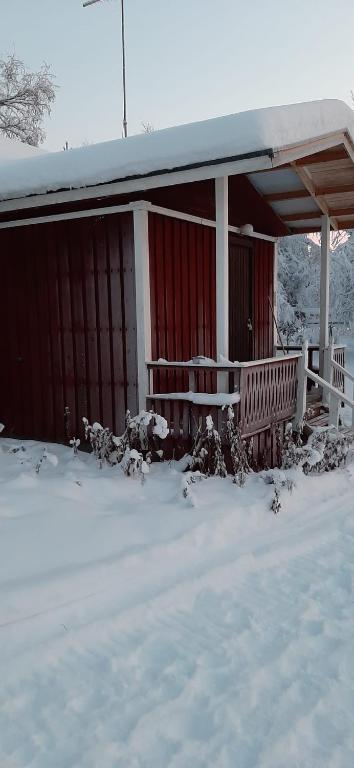 Pieni punainen puinen mökki, jossa on valkoiset reunukset, seisoo lumisessa maisemassa. Lumi peittää katon, portaat ja maan, ja kuistin kaiteen lähellä näkyy paljaita kasveja.