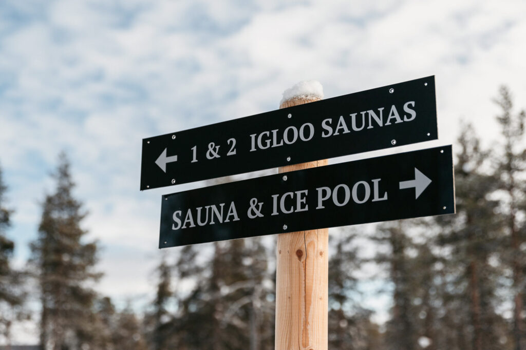 Puinen pylväs, jossa on kaksi mustaa opastekylttiä. Ylimmässä kyltissä lukee vasemmalle 1 & 2 Igloo Saunas. Alempi kyltti osoittaa oikealle ja siinä lukee Sauna & Ice Pool. Taustalla on puita ja pilvinen taivas.