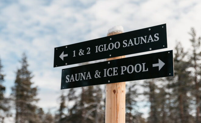 Puinen pylväs, jossa on kaksi mustaa opastekylttiä. Ylimmässä kyltissä lukee vasemmalle 1 & 2 Igloo Saunas. Alempi kyltti osoittaa oikealle ja siinä lukee Sauna & Ice Pool. Taustalla on puita ja pilvinen taivas.