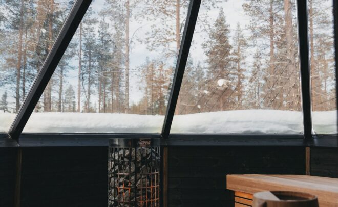 Viihtyisä sauna, jossa on puiset penkit ja metallinen kiuas, sijaitsee lasiseinäisessä huoneessa, josta avautuu selkeä näkymä lumipeitteiseen maahan ja korkeisiin mäntyihin ulkona sinisen taivaan alla.