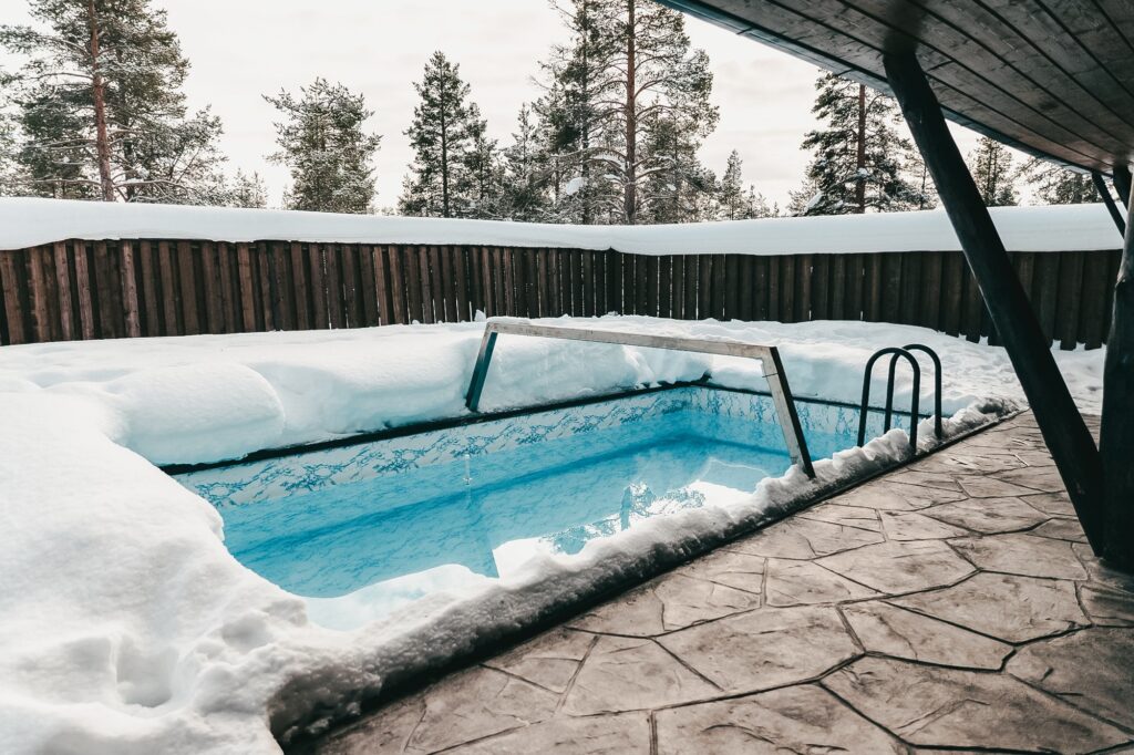 Syvän lumen ympäröimä uima-allas, taustalla puuaita ja mäntyjä. Uima-allasalue on osittain katettu katolla, ja maa on päällystetty kivilaatoilla.