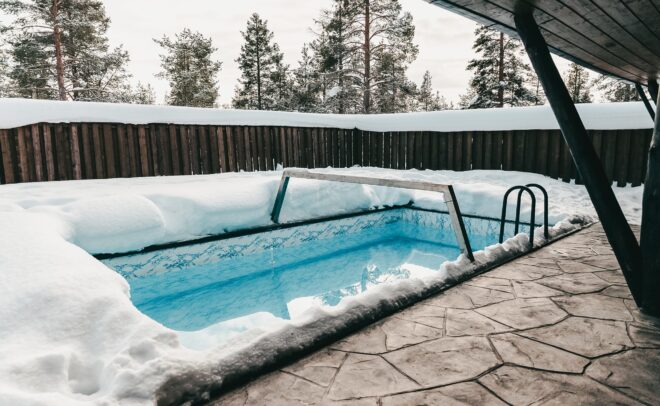 Syvän lumen ympäröimä uima-allas, taustalla puuaita ja mäntyjä. Uima-allasalue on osittain katettu katolla, ja maa on päällystetty kivilaatoilla.