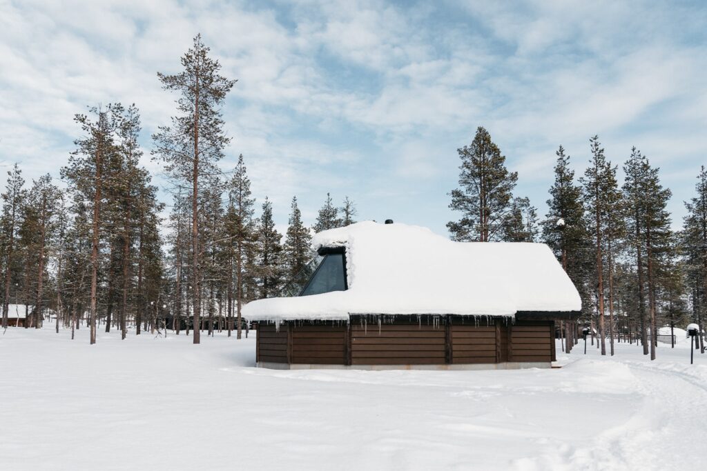 Pieni puinen mökki, jossa on suuri lumen peittämä katto ja jääpuikkoja, sijaitsee lumisessa metsässä, jota ympäröivät korkeat männyt osittain pilvisen taivaan alla.