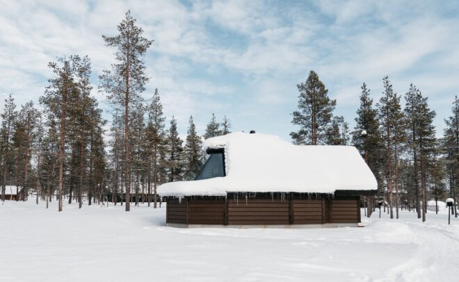 Pieni puinen mökki, jossa on suuri lumen peittämä katto ja jääpuikkoja, sijaitsee lumisessa metsässä, jota ympäröivät korkeat männyt osittain pilvisen taivaan alla.