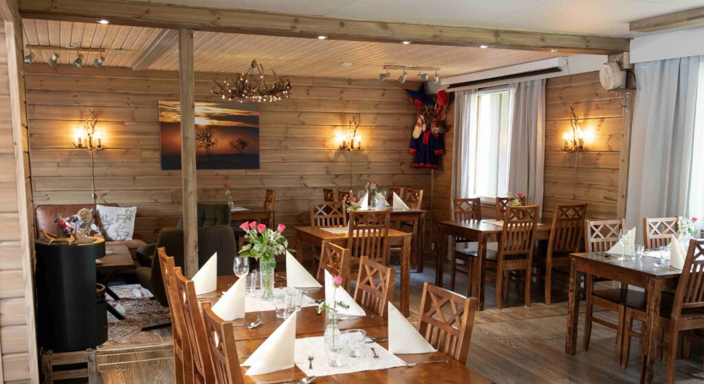 Viihtyisä ravintola, jossa on puiset seinät ja huonekalut, siististi katetut pöydät valkoisilla lautasliinoilla ja vaaleanpunaisilla ruusuilla maljakoissa, pehmeä valaistus ja lämmin, kutsuva tunnelma. Maalaus ja perinteiset vaatteet koristavat seiniä.
