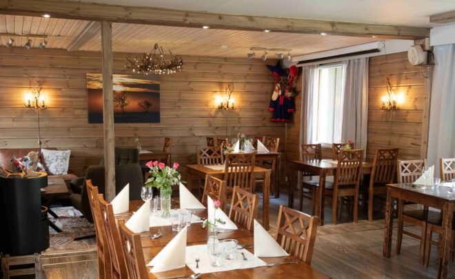 Viihtyisä ravintola, jossa on puiset seinät ja huonekalut, siististi katetut pöydät valkoisilla lautasliinoilla ja vaaleanpunaisilla ruusuilla maljakoissa, pehmeä valaistus ja lämmin, kutsuva tunnelma. Maalaus ja perinteiset vaatteet koristavat seiniä.