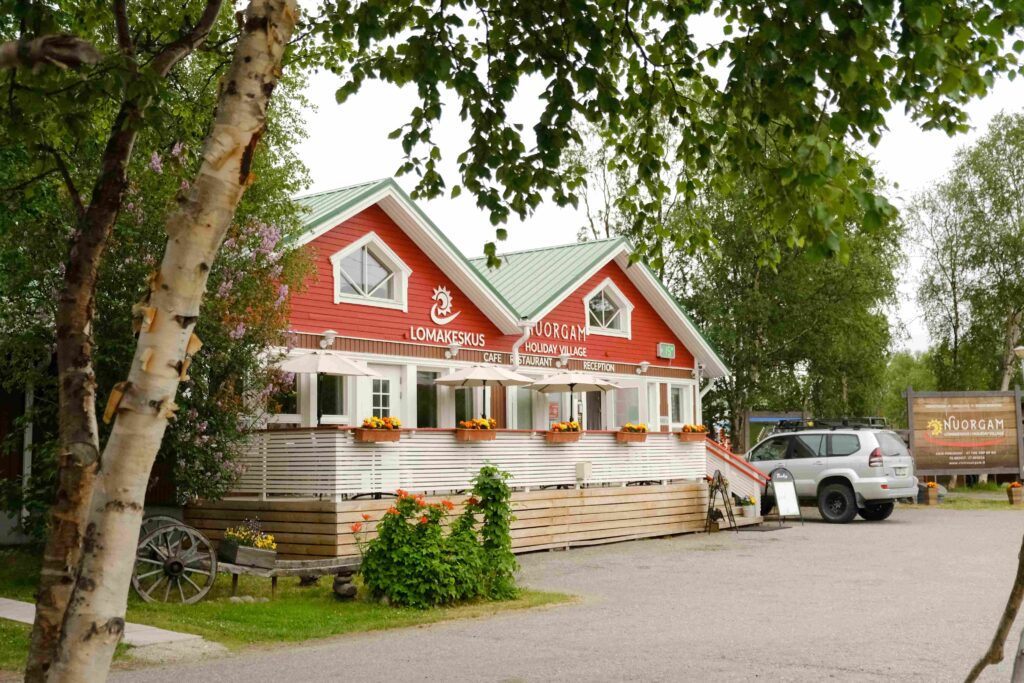 Punainen puurakennus, jossa on vihreä katto, valkoiset reunukset ja kuistilla kukkalaatikot. Auto on pysäköity lähelle, ja aluetta ympäröivät puut. Kyltissä lukee Nuorgamin lomakylä suomeksi ja englanniksi.