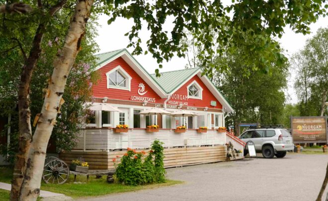 Punainen puurakennus, jossa on vihreä katto, valkoiset reunukset ja kuistilla kukkalaatikot. Auto on pysäköity lähelle, ja aluetta ympäröivät puut. Kyltissä lukee Nuorgamin lomakylä suomeksi ja englanniksi.