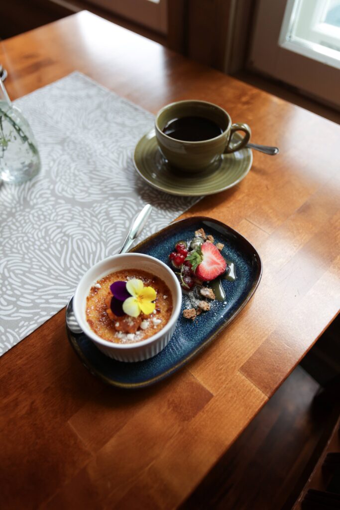 Kuppi mustaa kahvia istuu ikkunan vieressä olevalla puupöydällä lautasen vieressä, jossa on crème brûlée, jonka päällä on syötäviä kukkia, marjoja, tomusokeria ja mansikka.