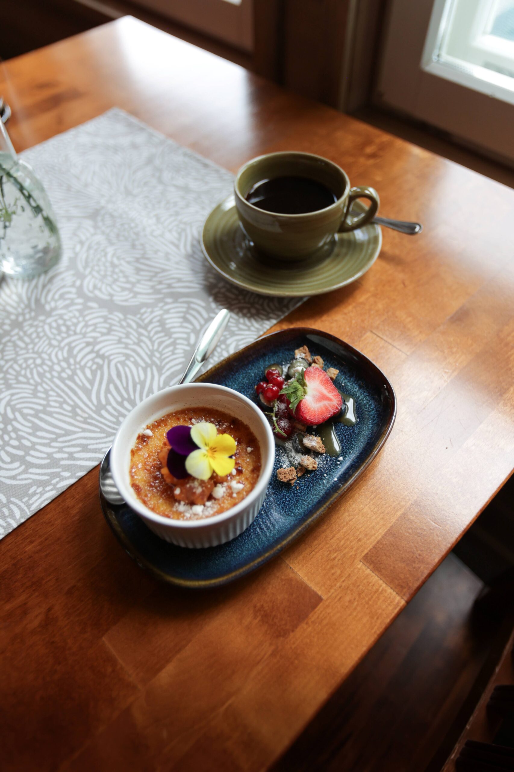 Kuppi mustaa kahvia istuu ikkunan vieressä olevalla puupöydällä lautasen vieressä, jossa on crème brûlée, jonka päällä on syötäviä kukkia, marjoja, tomusokeria ja mansikka.