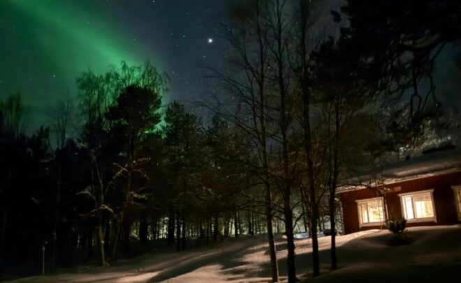Viihtyisä talo, jossa on lämpimästi valaistut ikkunat, sijaitsee lumisen polun varrella, puiden ympäröimänä ja vihreiden revontulien valaiseman tähtitaivaan alla.