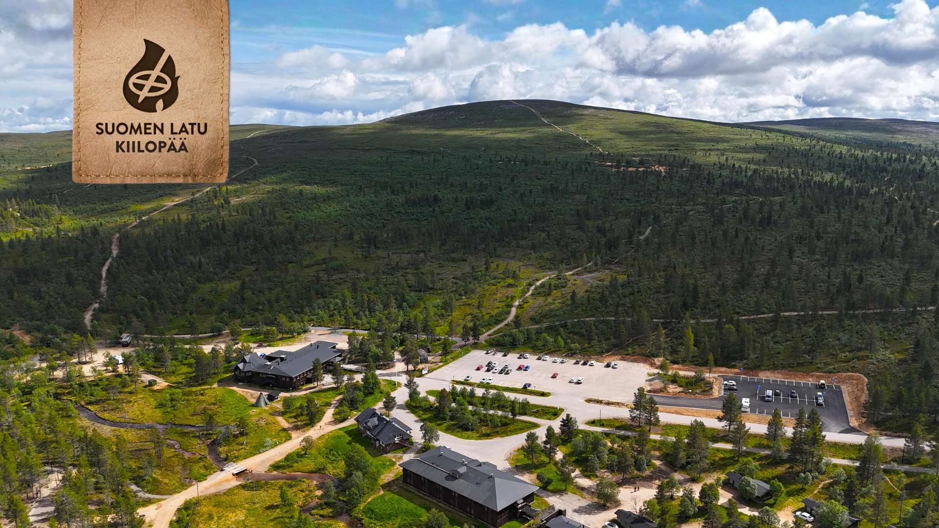 Ilmakuva metsäisestä vuoristoalueesta, jossa on useita rakennuksia, suuri parkkipaikka, jossa on autoja, ja mutkittelevia polkuja. Nurkassa olevassa puisessa kyltissä lukee Suomen Latu Kiilopää.