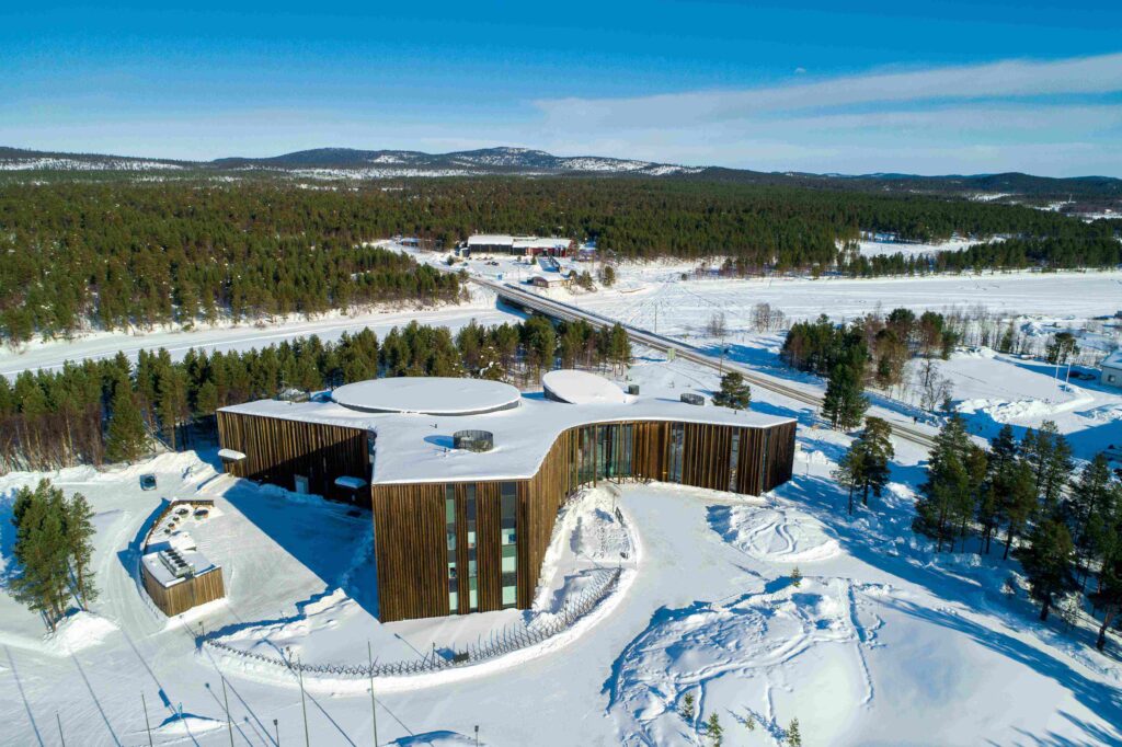 Ilmakuva modernista, kaarevasta puurakennuksesta lumen ja mäntyjen ympäröimänä maaseudulla vuoristomaisemassa kirkkaalla sinisellä taivaalla.