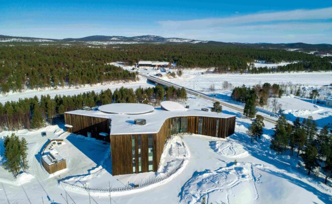 Ilmakuva modernista, kaarevasta puurakennuksesta lumen ja mäntyjen ympäröimänä maaseudulla vuoristomaisemassa kirkkaalla sinisellä taivaalla.