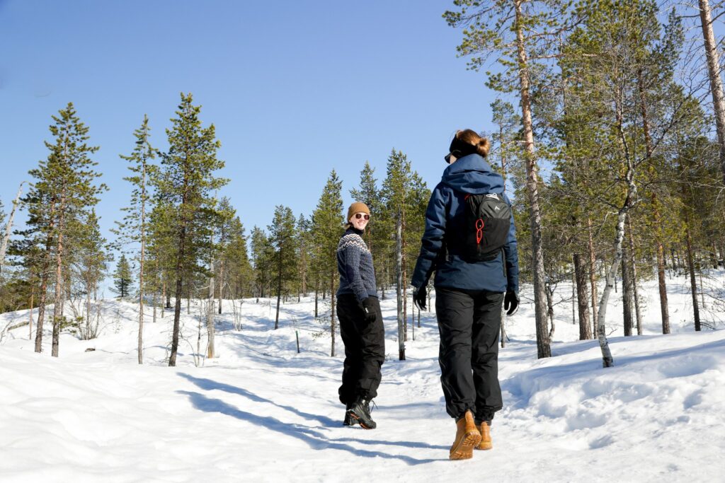 Kaksi talvivaatteisiin pukeutunutta ihmistä kävelee lumisessa metsässä kirkkaansinisen taivaan alla. Toinen henkilö katsoo taaksepäin ja hymyilee toiselle. Mäntyjä on hajallaan lumisessa maisemassa.