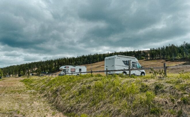 Kaksi matkailuautoa on pysäköity nurmikolle puuaidan lähelle; taustalla näkyy kumpuilevia kukkuloita ja mäntymetsää pilvisen taivaan alla.