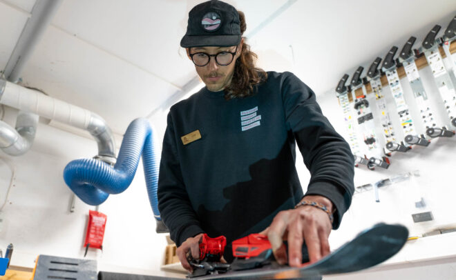 Silmälaseja ja hattua käyttävä henkilö säätää suksia työpajassa keskittyen tarkasti punaisen suksikiinnikkeen säätämiseen. Taustalla näkyy erilaisia työkaluja ja suksikiinnikkeitä.