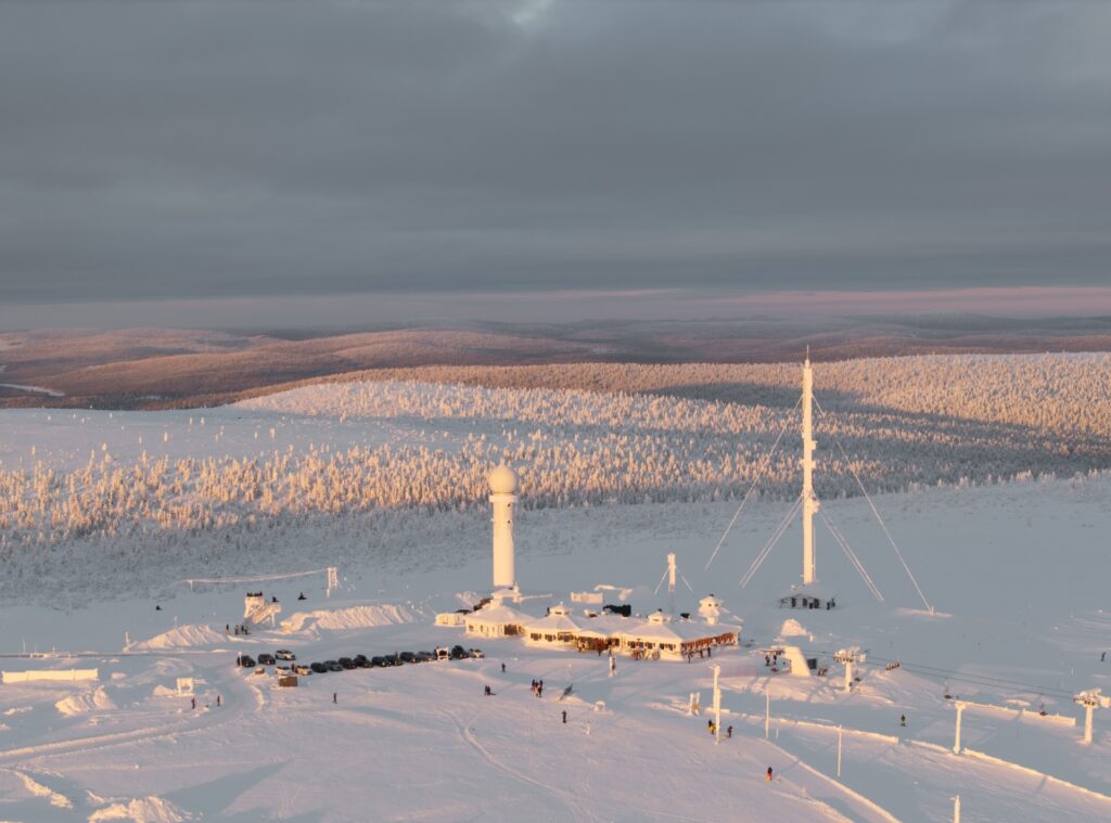 Luminen maisema, jossa on tieteellinen tutkimusasema, tutkakupolit ja korkea antenni. Aluetta ympäröivät lumen peittämät puut ja kukkulat pilvisen taivaan alla, ja pehmeä auringonvalo valaisee maisemaa.