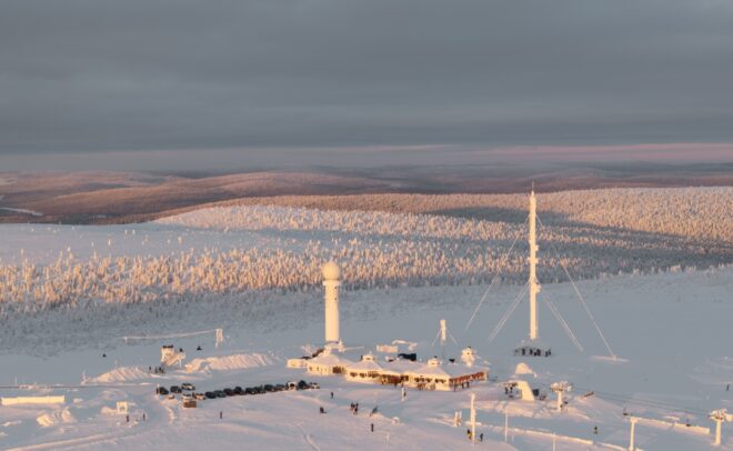 Luminen maisema, jossa on tieteellinen tutkimusasema, tutkakupolit ja korkea antenni. Aluetta ympäröivät lumen peittämät puut ja kukkulat pilvisen taivaan alla, ja pehmeä auringonvalo valaisee maisemaa.