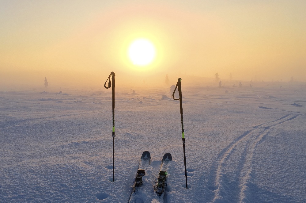 Pari suksia ja kaksi suksisauvaa seisovat pystyssä lumisessa maisemassa auringonnousun tai -laskun aikaan; suksijäljet jatkuvat kaukaisuuteen ja utuinen aurinko loistaa sumuisella taivaalla.