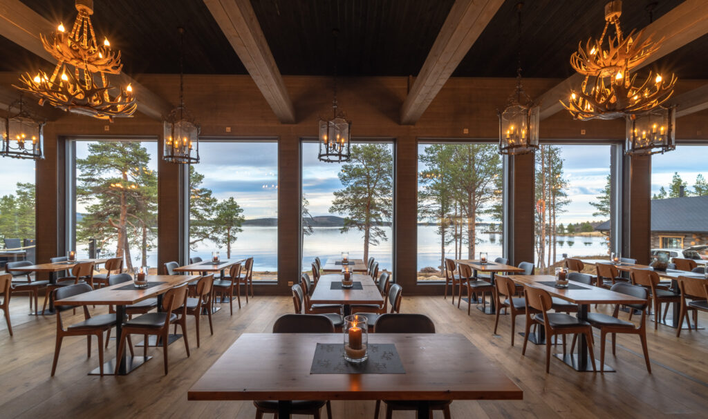 Viihtyisä ravintola, jossa on puupöytiä ja -tuoleja, sarvikruunut ja suuret ikkunat, joista avautuu näkymä järvelle ja mäntyihin auringonlaskun aikaan. Lämmin valaistus luo kutsuvan ilmapiirin.